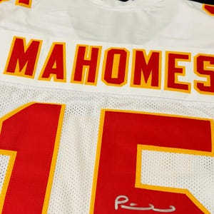 Puede incluir: Una camiseta deportiva de malla blanca con el nombre MAHOMES en letras rojas con bordes amarillos en la parte posterior. El n&uacute;mero 15 aparece en grandes d&iacute;gitos rojos con bordes amarillos, y una firma plateada es visible en el n&uacute;mero cinco.