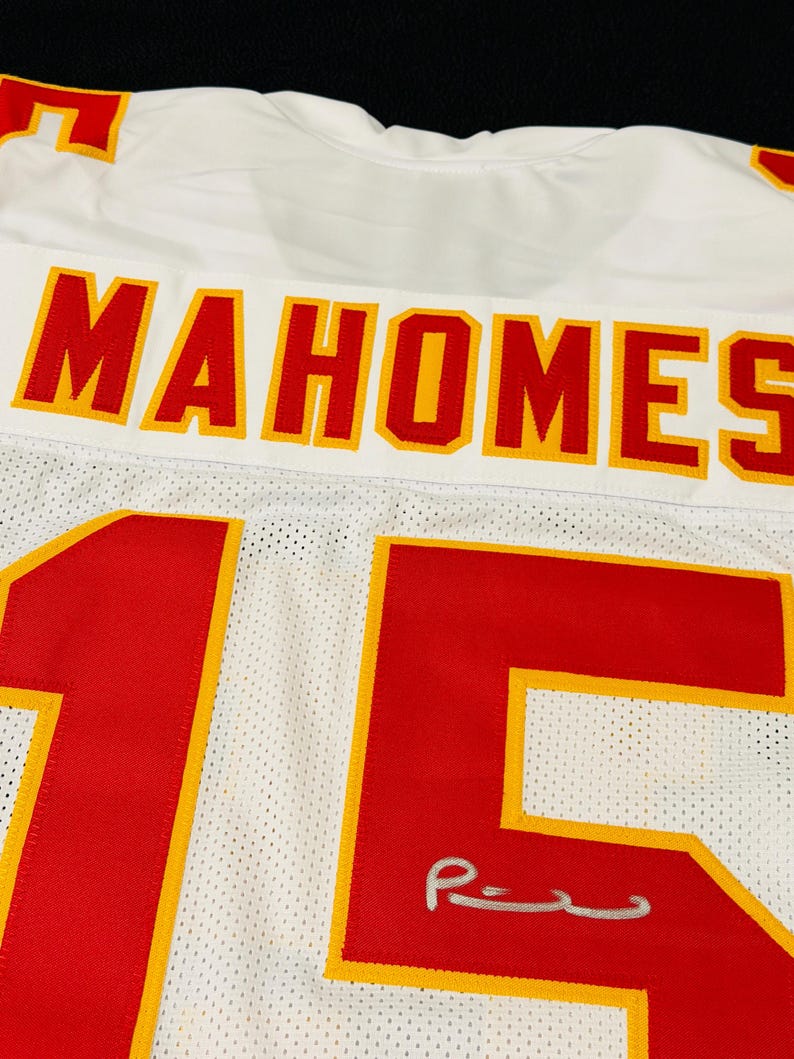 Puede incluir: Una camiseta deportiva de malla blanca con el nombre MAHOMES en letras rojas con bordes amarillos en la parte posterior. El n&uacute;mero 15 aparece en grandes d&iacute;gitos rojos con bordes amarillos, y una firma plateada es visible en el n&uacute;mero cinco.