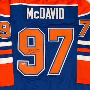 May include: A blue hockey jersey with orange and white accents. The jersey features the name "McDAVID" across the shoulders and the number "97" in orange with a white outline on the back. The jersey also has a signature.