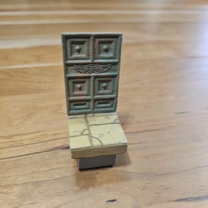 Op de afbeelding: Een miniatuur, verweerde deur met een vierkant paneelontwerp en een paar vleugels boven het midden. De deur is bevestigd aan een kleine, rechthoekige basis met een gebarsten, steenachtige textuur. Het kleurenschema is voornamelijk gedempt groen en bruin.