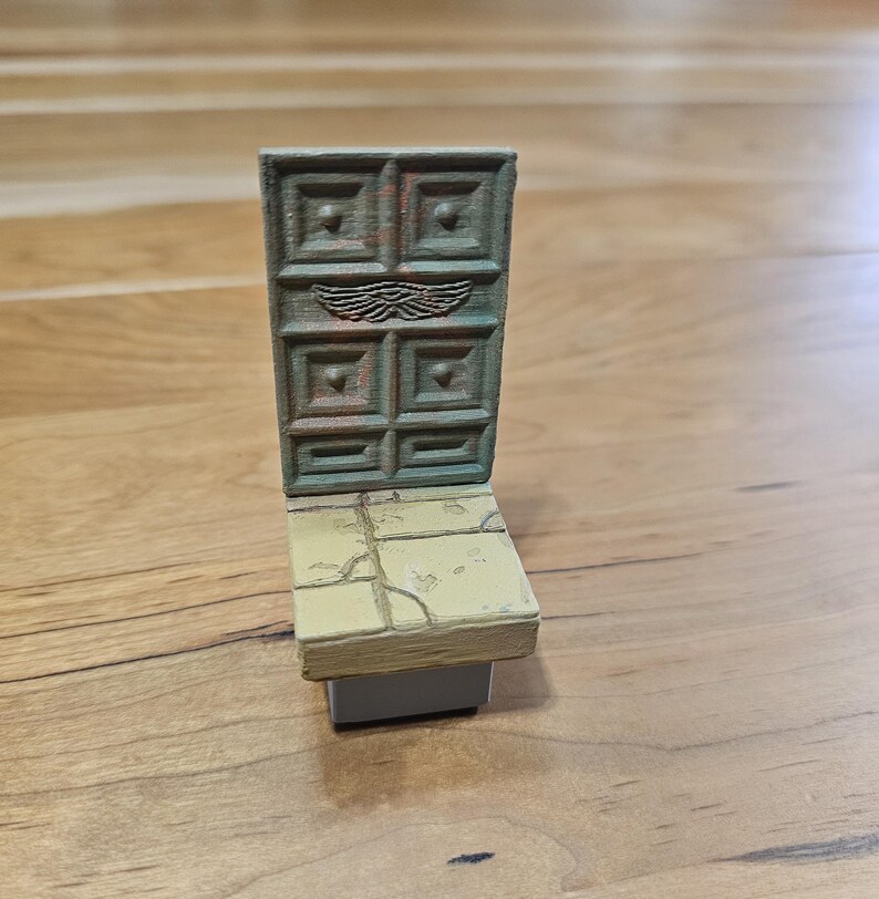 Op de afbeelding: Een miniatuur, verweerde deur met een vierkant paneelontwerp en een paar vleugels boven het midden. De deur is bevestigd aan een kleine, rechthoekige basis met een gebarsten, steenachtige textuur. Het kleurenschema is voornamelijk gedempt groen en bruin.