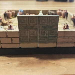 Op de afbeelding: Miniatuur diorama van een oude tempel of structuur, met beige metselwerk, decoratieve deuren en kleine beeldjes. De basis is zwart en het algehele ontwerp suggereert een historisch of fantasiethema.