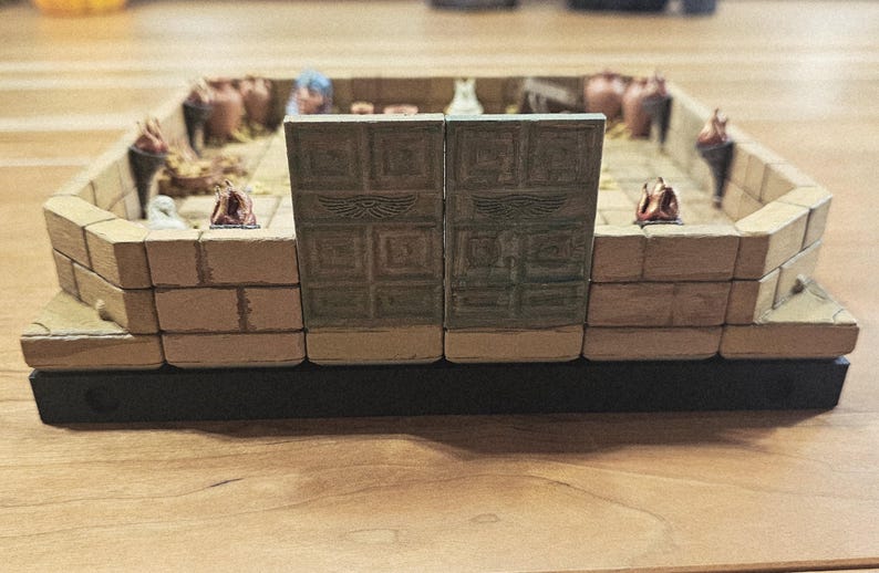 Op de afbeelding: Miniatuur diorama van een oude tempel of structuur, met beige metselwerk, decoratieve deuren en kleine beeldjes. De basis is zwart en het algehele ontwerp suggereert een historisch of fantasiethema.