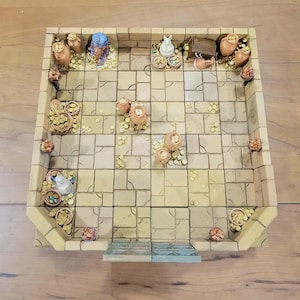 Op de afbeelding: Een miniatuur, rechthoekig kerker-spelbord met een steenachtig tegelpatroon. Het bord heeft kleine potten, schatkisten en beeldjes, waaronder een blauwe farao-buste en een witte kat. Gouden munten zijn verspreid.