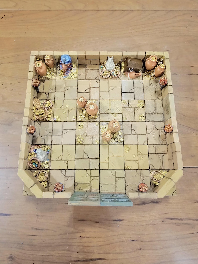 Op de afbeelding: Een miniatuur, rechthoekig kerker-spelbord met een steenachtig tegelpatroon. Het bord heeft kleine potten, schatkisten en beeldjes, waaronder een blauwe farao-buste en een witte kat. Gouden munten zijn verspreid.