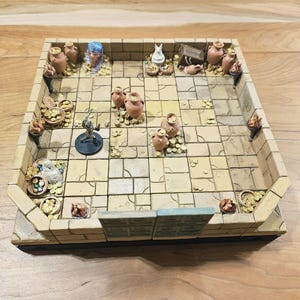 Op de afbeelding: Een miniatuur dungeon-spelbord met een steenachtige uitstraling. Het bord heeft een rasterpatroon, kleine kleipotten, gouden munten en een miniatuurfiguur. Het bord is rechthoekig met verhoogde randen.