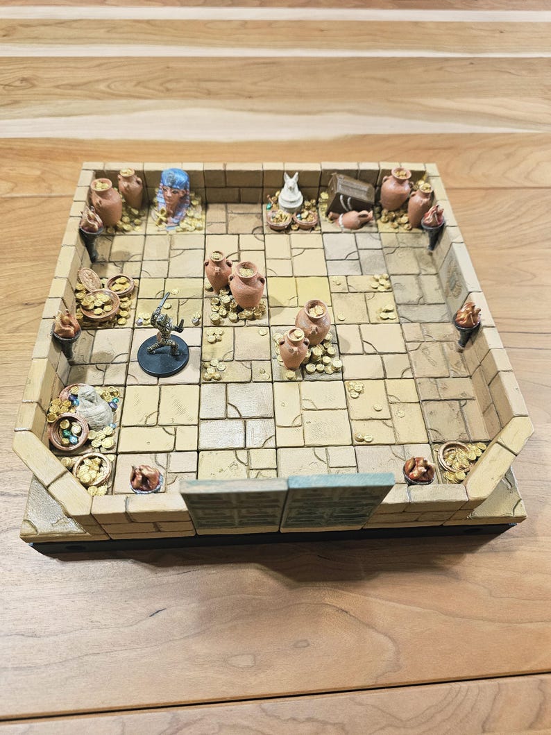 Op de afbeelding: Een miniatuur dungeon-spelbord met een steenachtige uitstraling. Het bord heeft een rasterpatroon, kleine kleipotten, gouden munten en een miniatuurfiguur. Het bord is rechthoekig met verhoogde randen.
