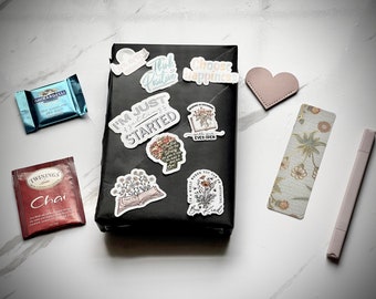 May include: A black wrapped gift box with various colorful stickers with motivational quotes like "I'm Just Getting Started", "Think Positive", "Choose Happiness", and "Be Kind". The box is surrounded by a tea bag, a chocolate bar, a heart-shaped bookmark, and a floral bookmark.