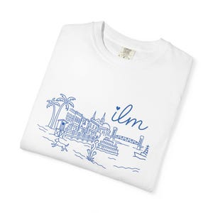 May include: White t-shirt with a blue line drawing of a waterfront scene, including buildings, a bridge, and a boat. The word "ilm" is written in cursive. The shirt is folded.