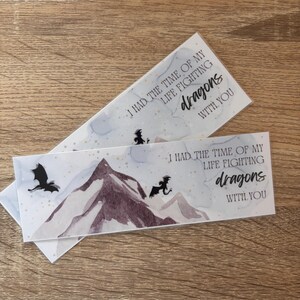 May include: Two rectangular bookmarks featuring a watercolour mountain scene and black dragon silhouettes. Each bookmark has the text "I HAD THE TIME OF MY LIFE FIGHTING dragons WITH YOU". The background has a light blue and grey watercolour effect.