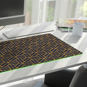 Puede incluir: Alfombrilla de ratón extendida con un patrón geométrico negro y dorado y un borde iluminado en verde. La alfombrilla está sobre un escritorio blanco con un monitor de ordenador, teclado y ratón. Un jarrón de flores está a la izquierda.