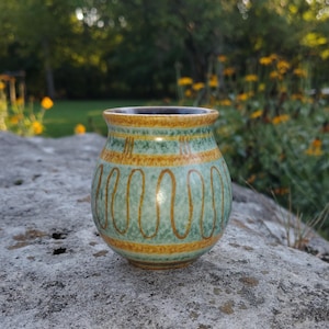 May include: A small, rounded ceramic cup featuring a mottled light green glaze with horizontal bands of mustard yellow. The exterior is decorated with a repeating vertical wavy line pattern in mustard yellow, and the interior rim is dark brown.