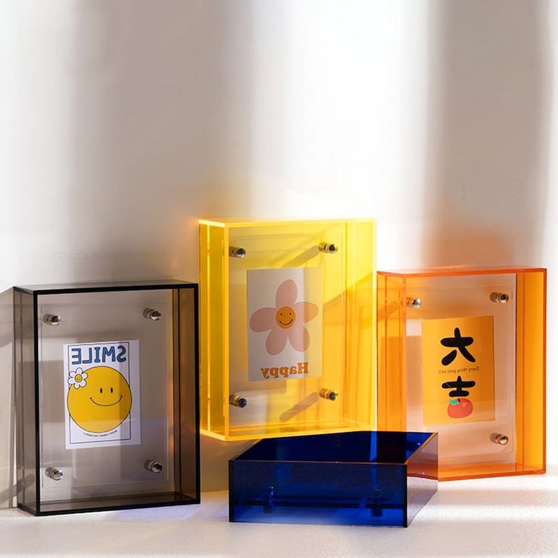 Op de afbeelding: Een verzameling kleurrijke acryl fotolijsten in verschillende maten en vormen. De lijsten zijn in tinten transparant, geel, oranje en blauw. Elke lijst bevat een decoratieve kaart met een grafisch ontwerp. De kaarten hebben een smiley, een bloem en Chinese karakters.