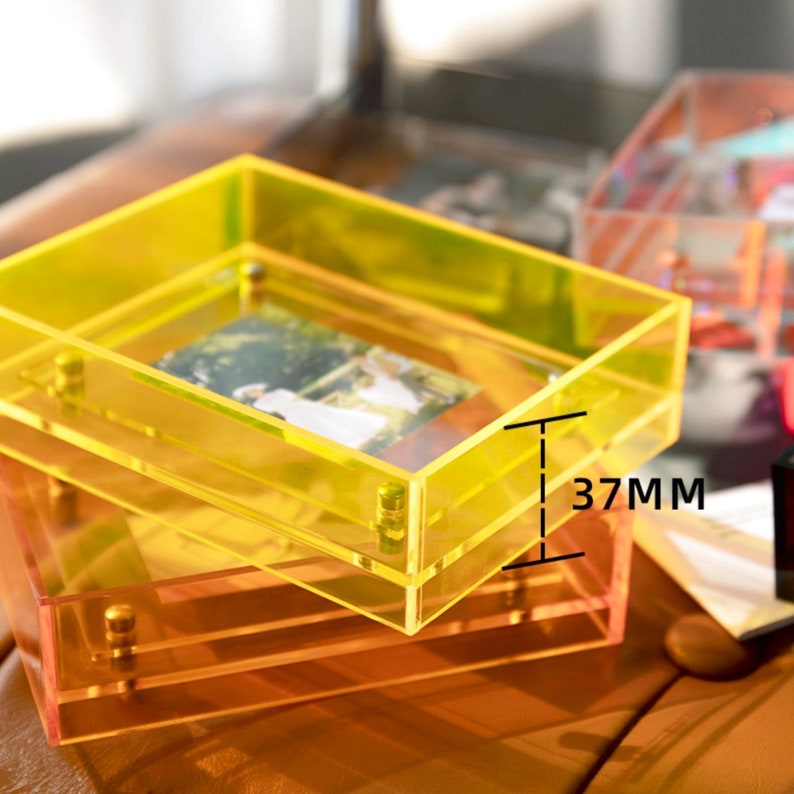 Op de afbeelding: Twee heldere acryl fotolijsten, &eacute;&eacute;n gele en &eacute;&eacute;n oranje, op elkaar gestapeld. De bovenste lijst bevat een foto. De lijsten zijn 3,7 cm dik en hebben goudkleurige hardware.