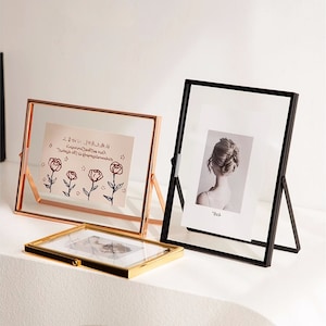 Op de afbeelding: Drie decoratieve fotolijsten in verschillende stijlen. Eén is een rechthoekige gouden lijst, een andere is een koperkleurige lijst met een standaard en de derde is een zwarte lijst met een standaard. De lijsten bevatten foto's en kunstwerken.