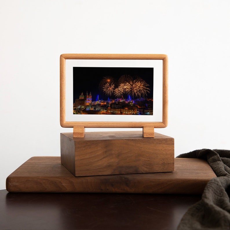 May include: A wooden picture frame with a stand displays a photograph of a city skyline at night with fireworks exploding in the sky.