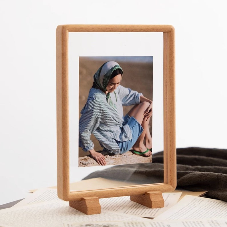 May include: A wooden picture frame with a stand, displaying a photo of a person wearing a green and white striped headscarf, a blue and white striped shirt, and blue denim shorts. The person is sitting on a sandy beach.