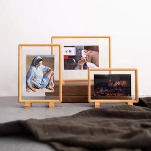 May include: Three wooden picture frames with different photos inside. The frames are standing on a brown surface. The photos show a woman sitting on the ground, a couple walking down a street, and fireworks over a city.