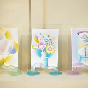 May include: Three glass picture frames with colorful abstract designs. The frames are on a white shelf and have a unique curved base.