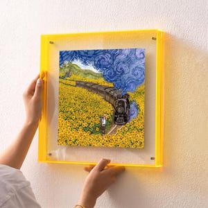 Op de afbeelding: Een vierkant wandkunstwerk met een kleurrijke illustratie van een stoomtrein die door een veld met gele zonnebloemen rijdt onder een wervelende blauwe lucht, gevat in een felgele, transparante acryl lijst met zilveren details.