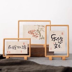 May include: Three wooden picture frames with clear glass fronts. The frames are standing on a light brown surface. The frames are different sizes and each has a different image inside. The smallest frame has Chinese characters that read "前程似锦". The middle frame has a watercolor painting of a branch with red berries. The largest frame has Chinese characters that read "乐 饮 文".