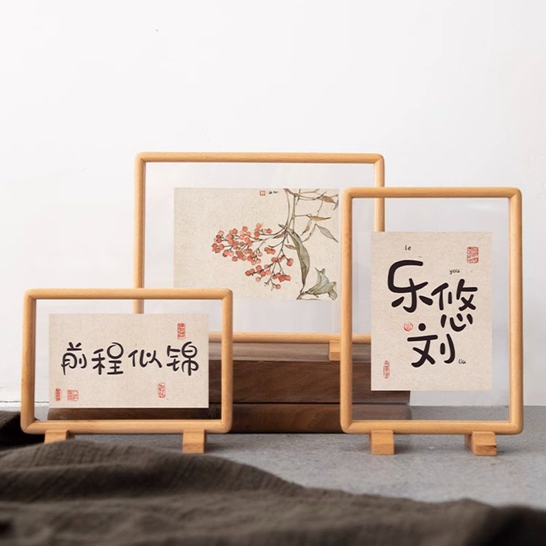 May include: Three wooden picture frames with clear glass fronts. The frames are standing on a light brown surface. The frames are different sizes and each has a different image inside. The smallest frame has Chinese characters that read "前程似锦". The middle frame has a watercolor painting of a branch with red berries. The largest frame has Chinese characters that read "乐 饮 文".