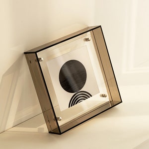 Puede incluir: Un marco de imagen de acr&iacute;lico transparente con una impresi&oacute;n geom&eacute;trica en blanco y negro en el interior. El marco tiene tornillos plateados en las esquinas.
