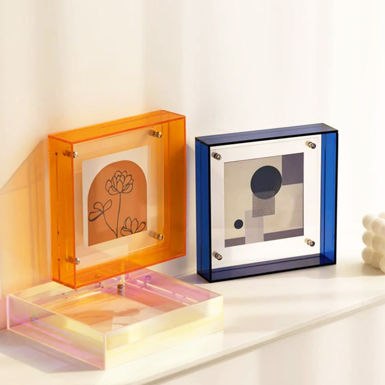 Puede incluir: Dos marcos de fotos de acr&iacute;lico con inserciones de vidrio transparente. El marco de la izquierda es naranja y el marco de la derecha es azul. Ambos marcos tienen un fondo blanco y un dise&ntilde;o simple en el interior.