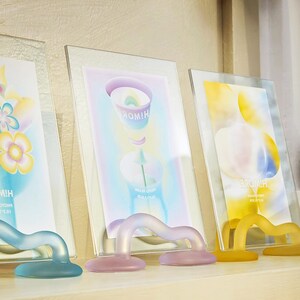 May include: Three glass picture frames with colorful abstract designs and the words "HIM!" and "HINMORE" printed on them. The frames are on a light wood surface and each has a clear plastic stand.