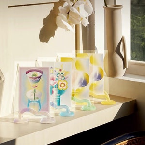 May include: Four colorful glass picture frames with abstract designs and the word "HIOMIH" printed on them. The frames are displayed on a white shelf with a beige vase in the background.