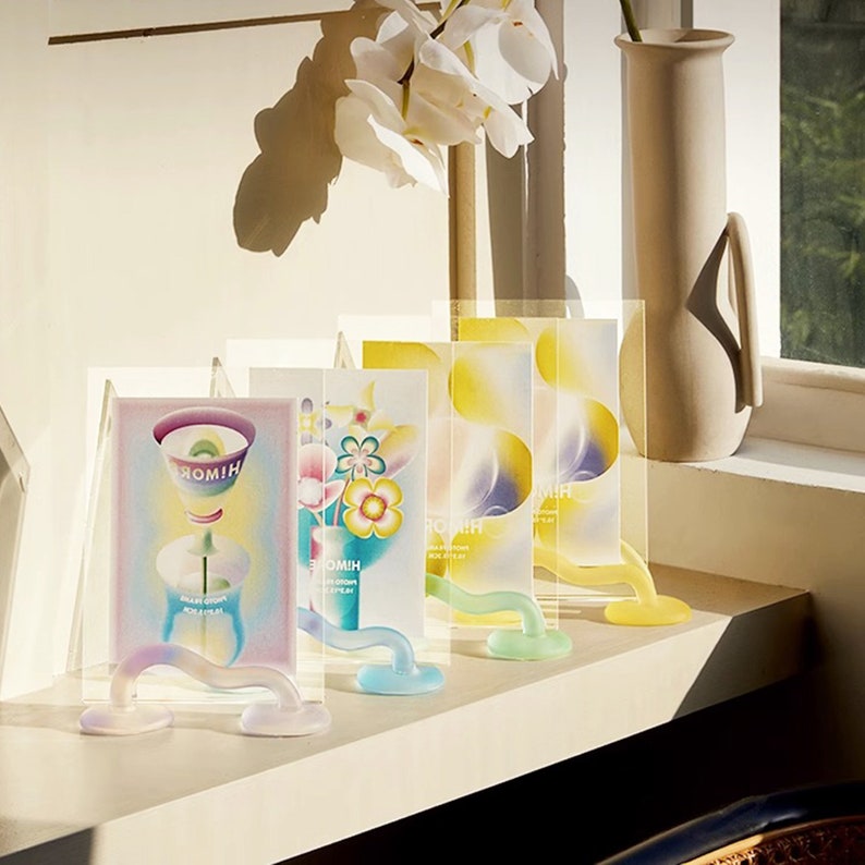 May include: Four colorful glass picture frames with abstract designs and the word "HIOMIH" printed on them. The frames are displayed on a white shelf with a beige vase in the background.