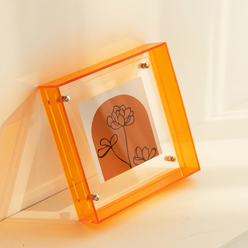 Puede incluir: Marco de fotos de acr&iacute;lico naranja con un estampado floral en el interior. El marco tiene un frente y una parte posterior de acr&iacute;lico transparente y se mantiene unido con tornillos plateados.