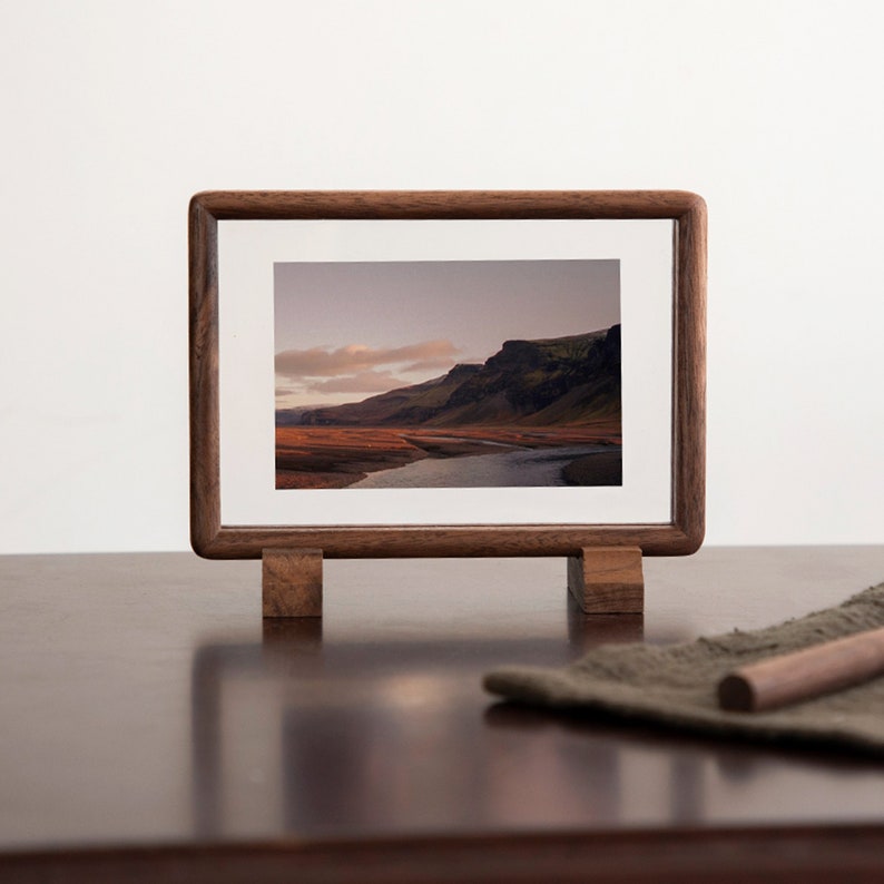 May include: A wooden picture frame with a landscape photograph of a river and mountains. The frame has a stand and is made of dark wood.