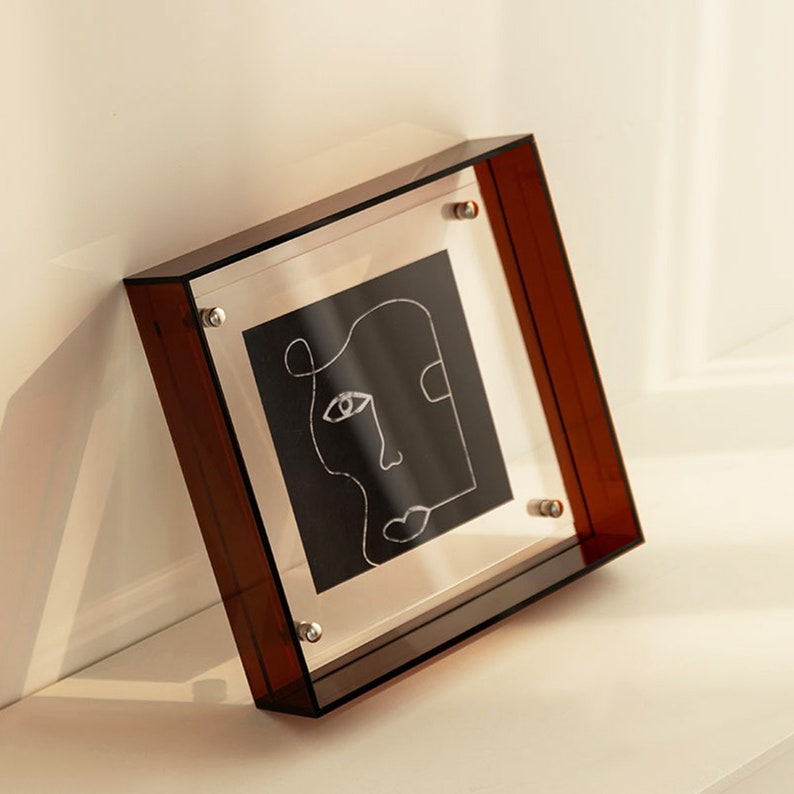 Puede incluir: Un marco de fotos de acr&iacute;lico marr&oacute;n con un dibujo de l&iacute;nea en blanco y negro de una cara en el interior. El marco tiene tornillos plateados en las esquinas.