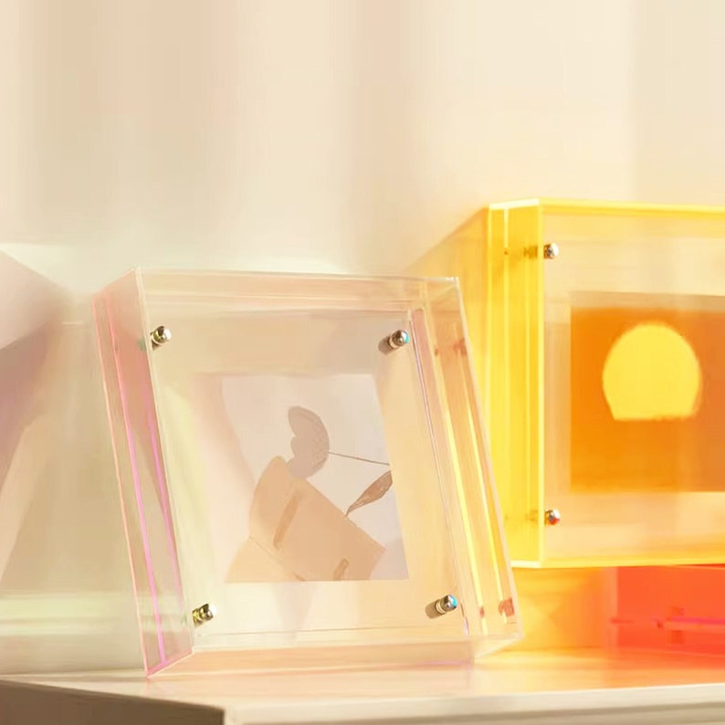 Puede incluir: Tres marcos de fotos de acr&iacute;lico transparente con detalles dorados. Los marcos son de diferentes colores: rosa, amarillo y naranja. Los marcos est&aacute;n vac&iacute;os, pero uno tiene una foto dentro.