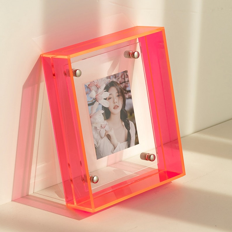 Op de afbeelding: Een neonroze acryl fotolijst met een helder midden, met een foto van een vrouw met bloemen. De vierkante lijst heeft zilverkleurige bevestigingen in de hoeken. De lijst staat tegen een witte ondergrond.
