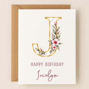 May include: A white greeting card featuring a large letter J with a white marble texture and gold trim, decorated with pink flowers and green leaves. Below the letter, the text reads HAPPY BIRTHDAY and the name Jocelyn in a pink script font.