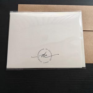 May include: A stack of off-white cards in a clear plastic sleeve, with a brown paper envelope. The cards have a textured surface and a circular logo that reads "HANDMADE BY DESIGNALIZE CRAFT" with the initials "dc".