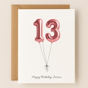 May include: A white greeting card featuring two metallic rose gold foil balloons shaped like the number 13, tied together with thin black strings. Below the balloons, the text Happy Birthday, Jessica is printed in a simple black script font.
