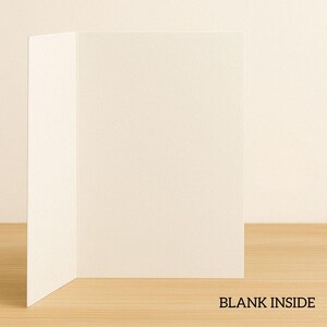 May include: Blank white card with a blank inside. The card is folded in half and is sitting on a light brown wooden surface.