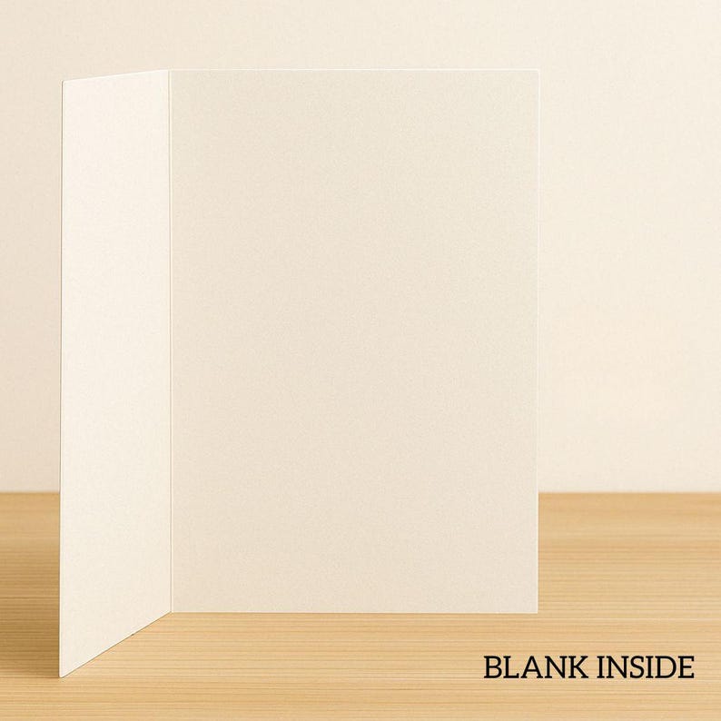 May include: Blank white card with a blank inside. The card is folded in half and is sitting on a light brown wooden surface.