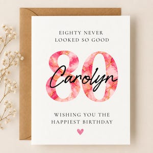 May include: A white birthday greeting card featuring the text EIGHTY NEVER LOOKED SO GOOD at the top, a large number 80 with a pink and gold marble pattern, the name Carolyn in black script, and WISHING YOU THE HAPPIEST BIRTHDAY with a small pink heart.