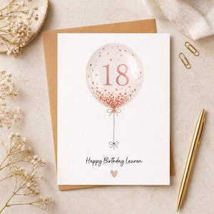 May include: A white greeting card featuring a central illustration of a translucent balloon filled with rose gold confetti and the number 18 in rose gold. Below the balloon, the text reads Happy Birthday Lauren with a small rose gold heart icon.