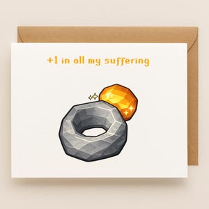 May include: Greeting card with a brown envelope. The card features a gray ring and a golden gem with the text "+1 in all my suffering" in orange. The design has a fantasy game aesthetic.