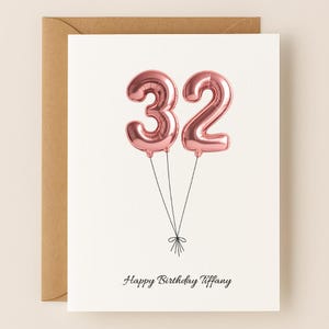May include: A birthday card with a white background and a brown envelope. The card features two rose gold number 32 balloons tied together with black string. The words "Happy Birthday Tiffany" are written in black script below the balloons.