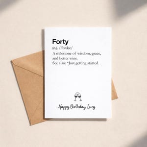 May include: A white birthday card with the word "Forty" at the top, followed by a definition. The card includes the text "Happy Birthday, Lucy" and two wine glasses. A brown envelope is behind the card.