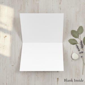 May include: An open, blank white greeting card on a light wood-grain surface. The card is centered, revealing a clean interior. To the right, sprigs of greenery and a small stone add a natural touch. The text "Blank Inside" is at the bottom.