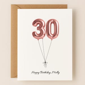 May include: A birthday card with a kraft paper envelope. The card features two rose gold foil number 30 balloons tied together with black string. The words "Happy Birthday, Molly" are written in a delicate script below the balloons.