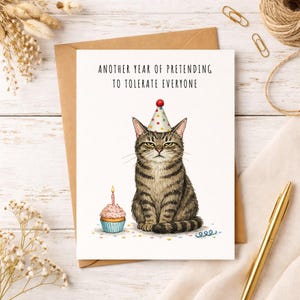 May include: A greeting card with a grumpy cat wearing a party hat and a cupcake with a lit candle. The card reads "ANOTHER YEAR OF PRETENDING TO TOLERATE EVERYONE". The cat is brown and gray with a serious expression.