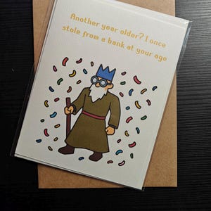 May include: A birthday card with a cartoon illustration of a man wearing a crown and glasses, holding a cane. The card reads "Another year older? I once stole from a bank at your age".